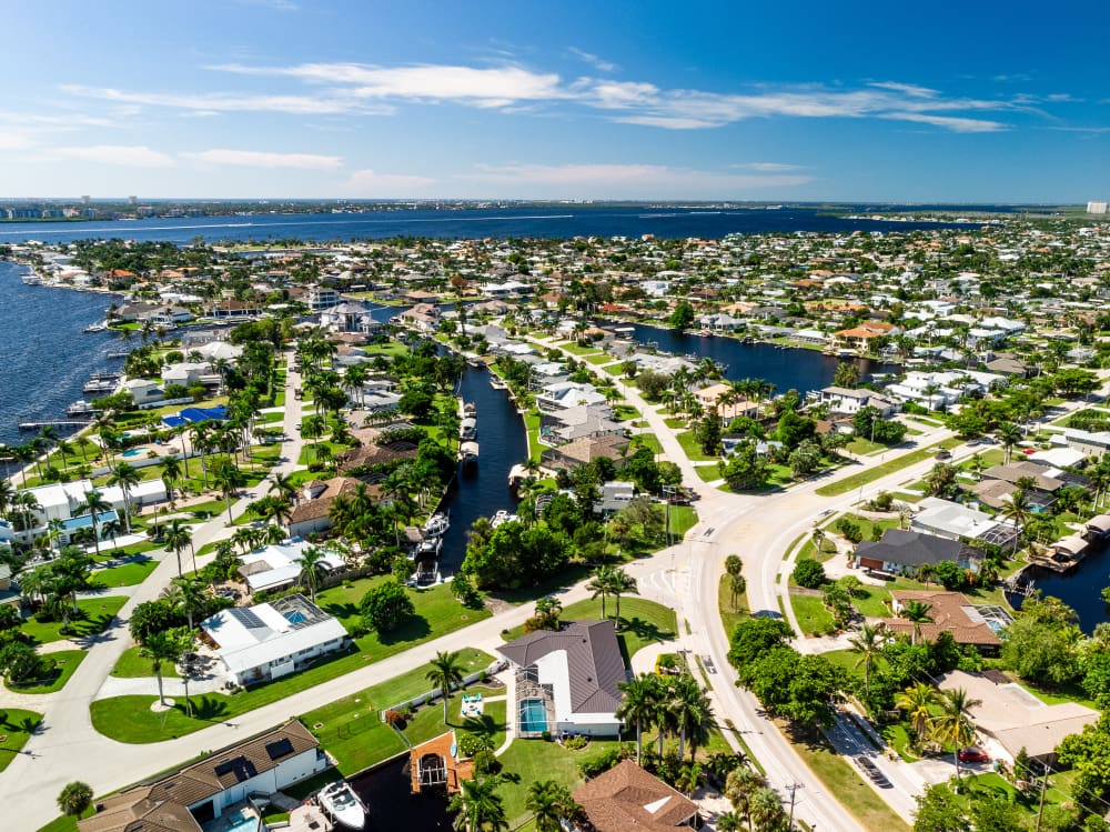An aerial view shows a sunny waterfront neighborhood with palm-lined streets, canals winding between houses, boats docked at homes, and wide blue water stretching toward the horizon.