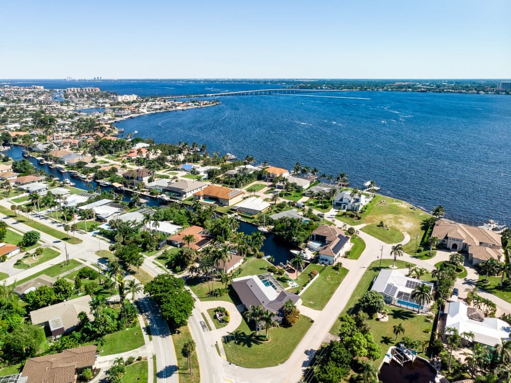 An aerial view reveals a waterfront neighborhood with winding canals, palm-lined streets, and houses with docks, opening out to wide blue water under a clear sky.