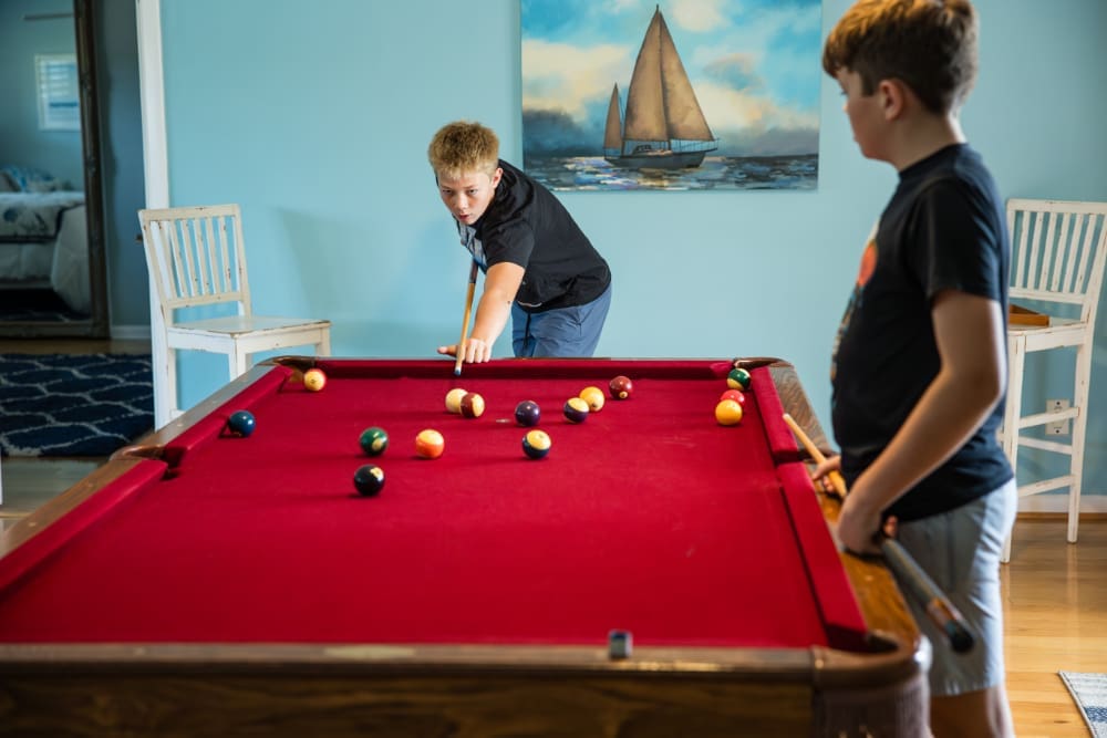 Two children play pool indoors at a red felt table, one lining up a shot while the other watches. Soft light and coastal décor give the room a relaxed vacation feel.