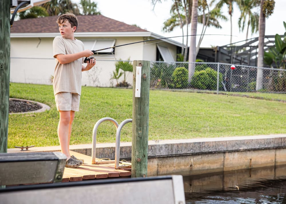 A young child stands on a small dock, casting a fishing line into a quiet canal. Green lawns, palm trees, and a neighborhood home create a peaceful backyard scene.