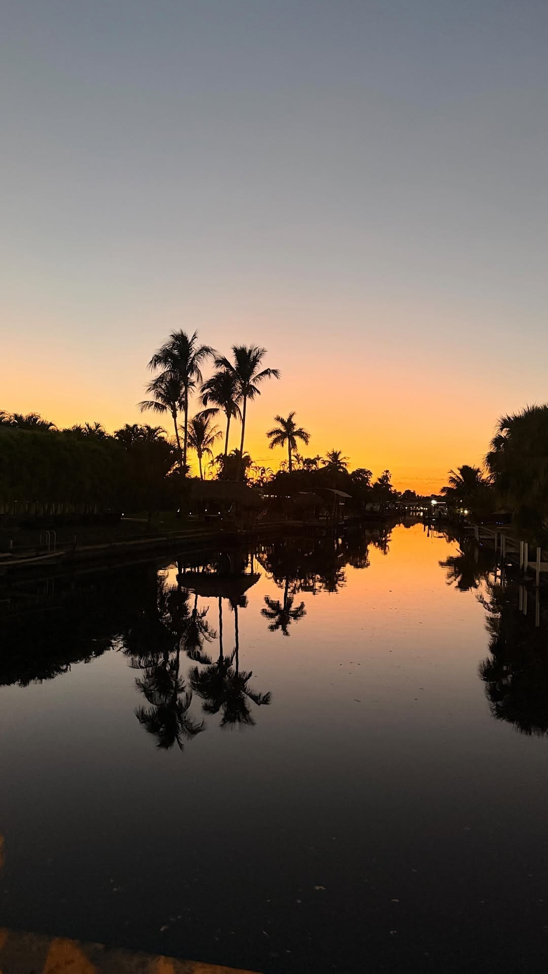 Sunset over calm canal with palm trees reflected in still water, creating a warm, peaceful view