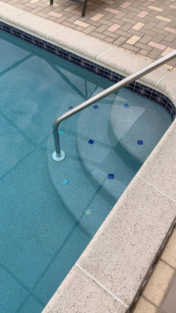 Clear pool water with entry steps and handrail, sunlit patio edge, and clean, inviting design